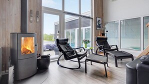A modern living room with a wood-burning stove, leather chairs, a small table, and large windows offering a view of the outdoors. - Awesome home in Ebeltoft with sauna (Ebeltoft)