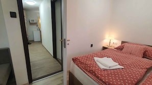 Ett hotellrum med en säng, ett sängbord med en lampa, en stol och utsikt över korridoren och det lilla köket.