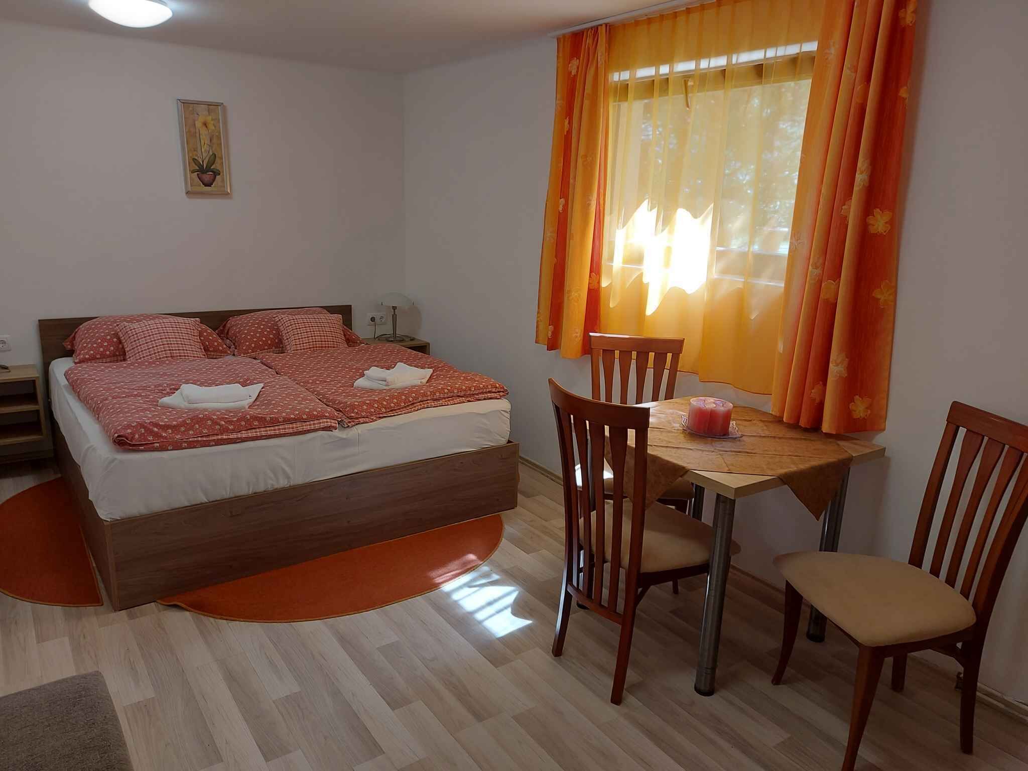 A bedroom with a bed, a wooden table with chairs, and a window with orange curtains.