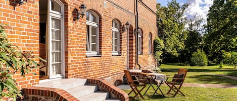 Een bakstenen gebouw met een stenen trap naar een deur, een tafel met stoelen en een tuin op de achtergrond.