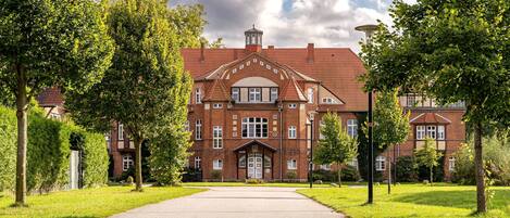 Ein großes Backsteinhaus mit einem zentralen Eingang, umgeben von Bäumen und einem gepflasterten Weg.
