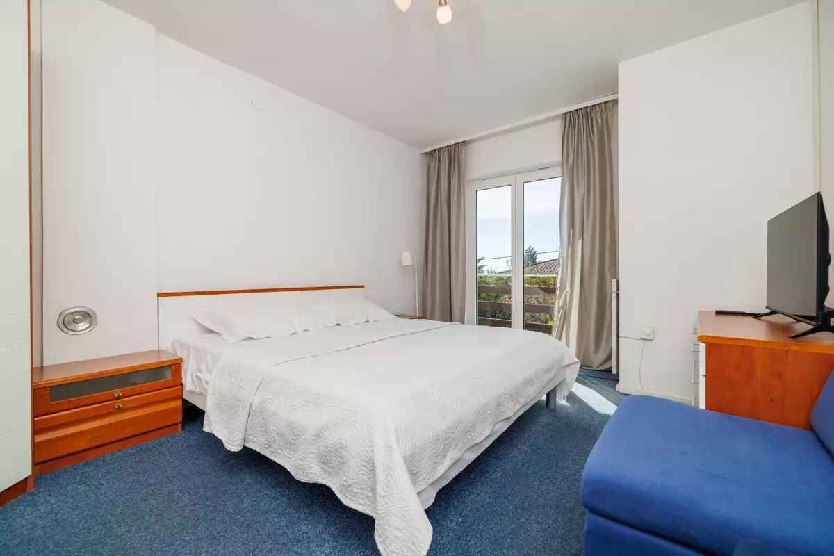 Een hotelkamer met een enkel bed, een houten nachtkastje, een blauwe fauteuil, een televisie en een groot raam met gordijnen.
