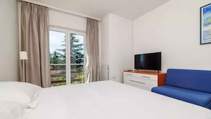 Une chambre d’hôtel avec un grand lit, un téléviseur posé sur une commode en bois, un canapé bleu et un balcon avec des rideaux.