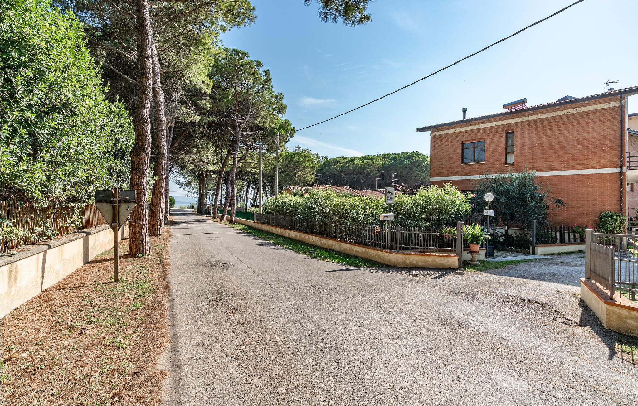 A quiet residential street with a gravel road, a brick building, and a fence with greenery.