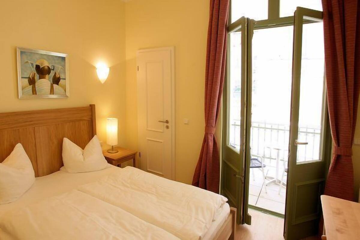 Quarto de hotel com cabeceira de madeira, cama com roupa de cama branca, duas luminárias de apoio, um quadro na parede e uma porta que dá para uma varanda.