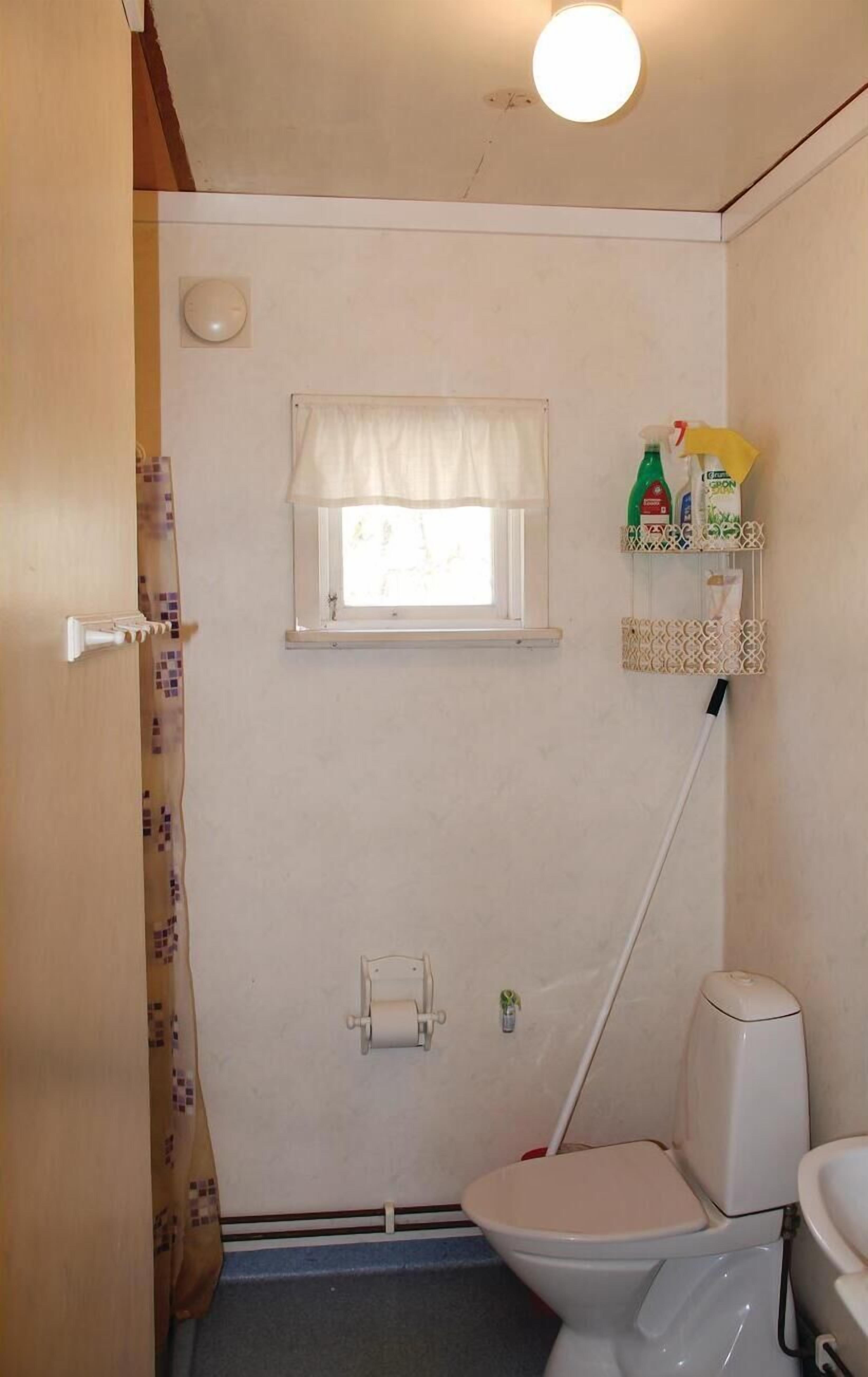 Een kleine, compacte badkamer met een toilet, een raam met een witte vouwgordijn, een toiletrolhouder aan de muur en een hangende toiletborstel.