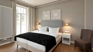 Moderni hotellihuone, jossa on suuri sänky, kaksi yöpöytää, tuoli ja koristeellinen seinätaideteos.