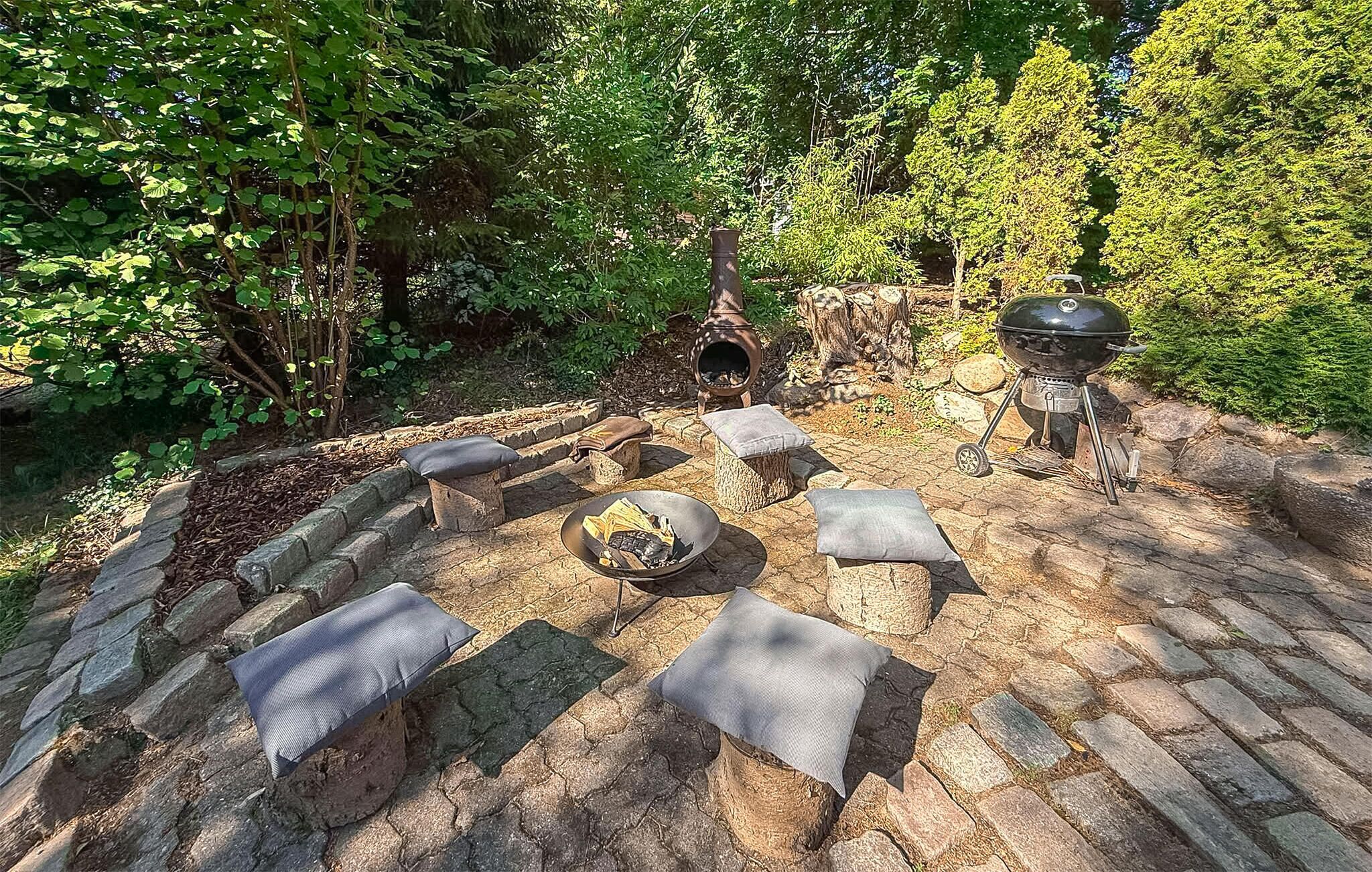 A stone patio with outdoor seating, a wood-fired oven, and a grill, surrounded by lush greenery.