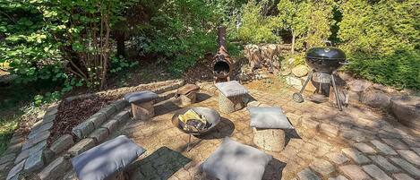 A stone patio with outdoor seating, a wood-fired oven, and a grill, surrounded by lush greenery.