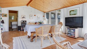 Een gezellige woonkamer met een houten plafond, een eettafel met rieten stoelen, een flatscreen televisie en een boekenkast.