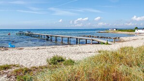 Een strand met een houten pier die zich uitstrekt in de zee, een klein gebouw aan de kust, en een heldere lucht.