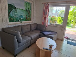 Un salón con un sofá gris, una mesa de centro redonda de madera, un cuadro de un paisaje y una ventana con cortina.