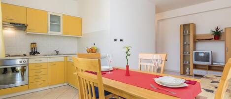 A compact kitchen with yellow cabinets, a dining table with a red tablecloth, and a TV in the living area.