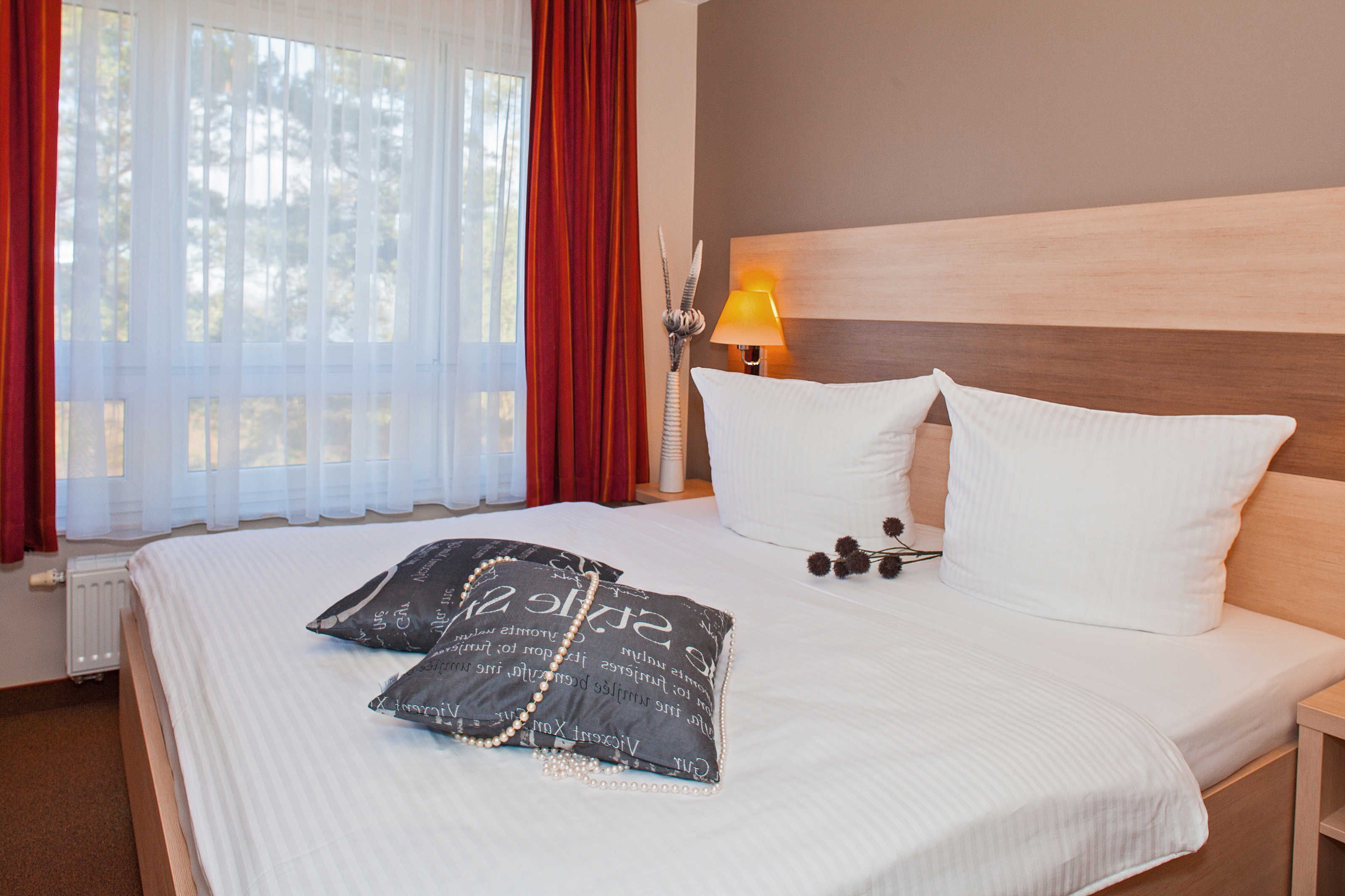 A neatly made bed with white and black pillows, a bedside lamp, and a window with red curtains.