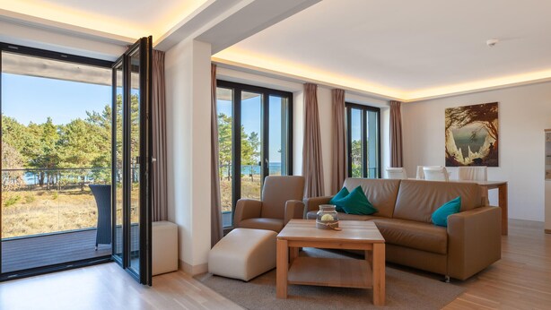 A modern living room with a sofa, ottoman, and coffee table, large windows with curtains, and a painting on the wall. - Gorgeous apartment in Binz with indoor swimming pool (Prora/Rügen)