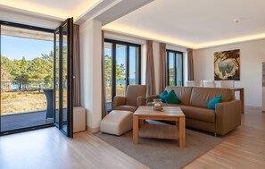 Una sala moderna con sofá, reposapiés y mesa de centro, ventanas amplias con cortinas y un cuadro en la pared.