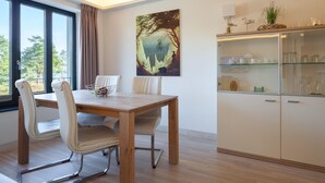 A dining area with a wooden table and chairs, a glass cabinet with kitchenware, a painting on the wall, and a view through the window. - Gorgeous apartment in Binz with indoor swimming pool (Prora/Rügen)