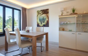 A dining area with a wooden table and chairs, a glass cabinet with kitchenware, a painting on the wall, and a view through the window.