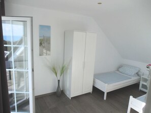 Un dormitorio con cama, armario, mesita de noche, jarra con planta y ventana con vistas.