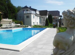 Una piscina con una estatua en primer plano, una casa con balcón al fondo y un cielo despejado.