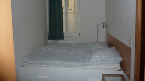 Una habitación pequeña con una cama individual, un colchón blanco sencillo, cabecero de madera, una mesita de noche, una lámpara de pie y una ventana con cortina de encaje.