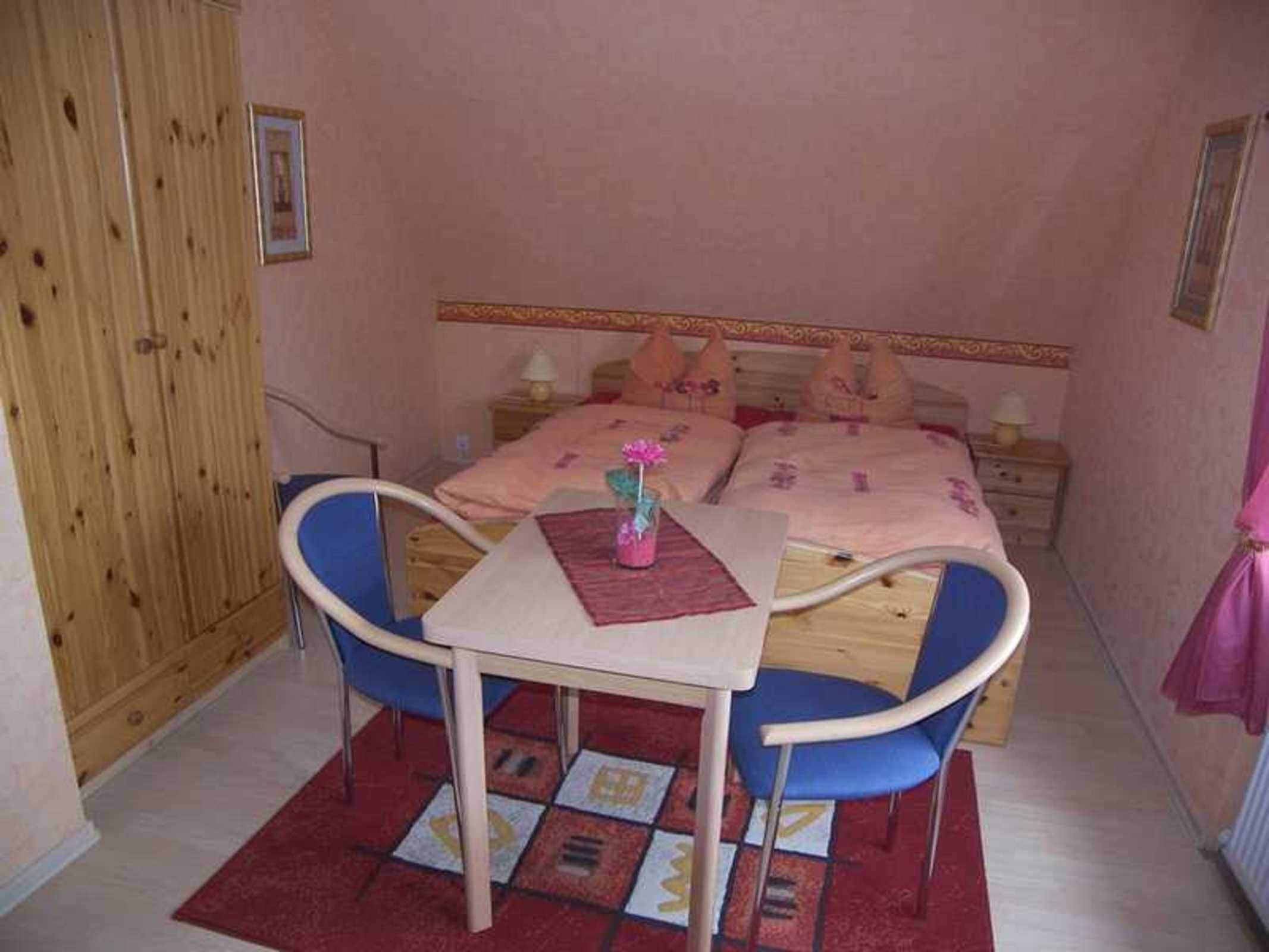 Un dormitorio con una cama, dos mesitas de noche, un armario de madera, una pequeña mesa con una silla y una alfombra roja.