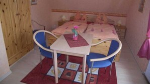 A bedroom with a bed, two bedside tables, a wooden wardrobe, a small table with a chair, and a red rug. - Apartment 02 - Apartments Oechsner (Heringsdorf)