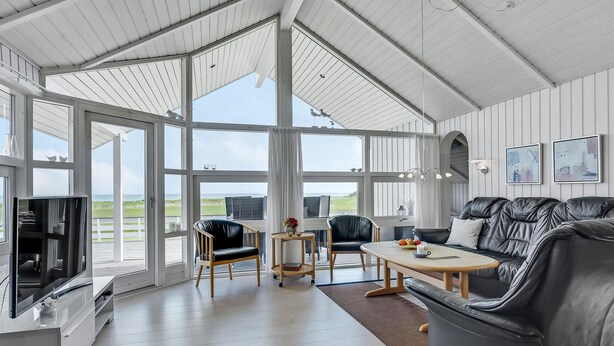 Een ruime kamer met een grote glazen deur die leidt naar een balkon, een zwarte leren bank, een houten salontafel en een televisie op een witte kast.