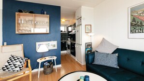 Ein kompakter Wohnbereich mit Küchenzeile, einer Couch mit Kissen, einem kleinen Tisch mit Lampe und einem gerahmten Bild an der Wand.