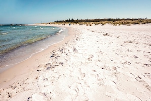 A sandy beach with clear water, a few footprints, and some dunes in the background.
