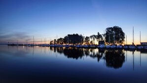 A marina at dusk with calm waters reflecting the boats and trees.