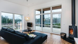 A modern living room with a black leather sofa, a TV on a white stand, a round table with a glass, and a large window overlooking a forest. - Beautiful home in Holmsjö with WiFi (Holmsjö)