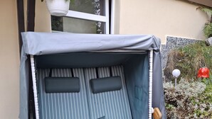 Eine hölzerne Bank mit Streifenpolstern und einem Kissen, draußen platziert, mit Fenster und Topfpflanzen in der Nähe.