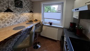 Eine kompakte Küche mit Holzarbeitsplatte, zwei Barhocker, einem Fenster mit Jalousien, einem Heizkörper und einem Herd.