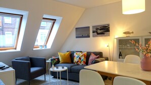 Una sala de estar moderna con un sofá, sillas, una mesita y una mesa de comedor. Hay dos ventanas con persianas, una lámpara de techo y un aparador con puertas de vidrio.