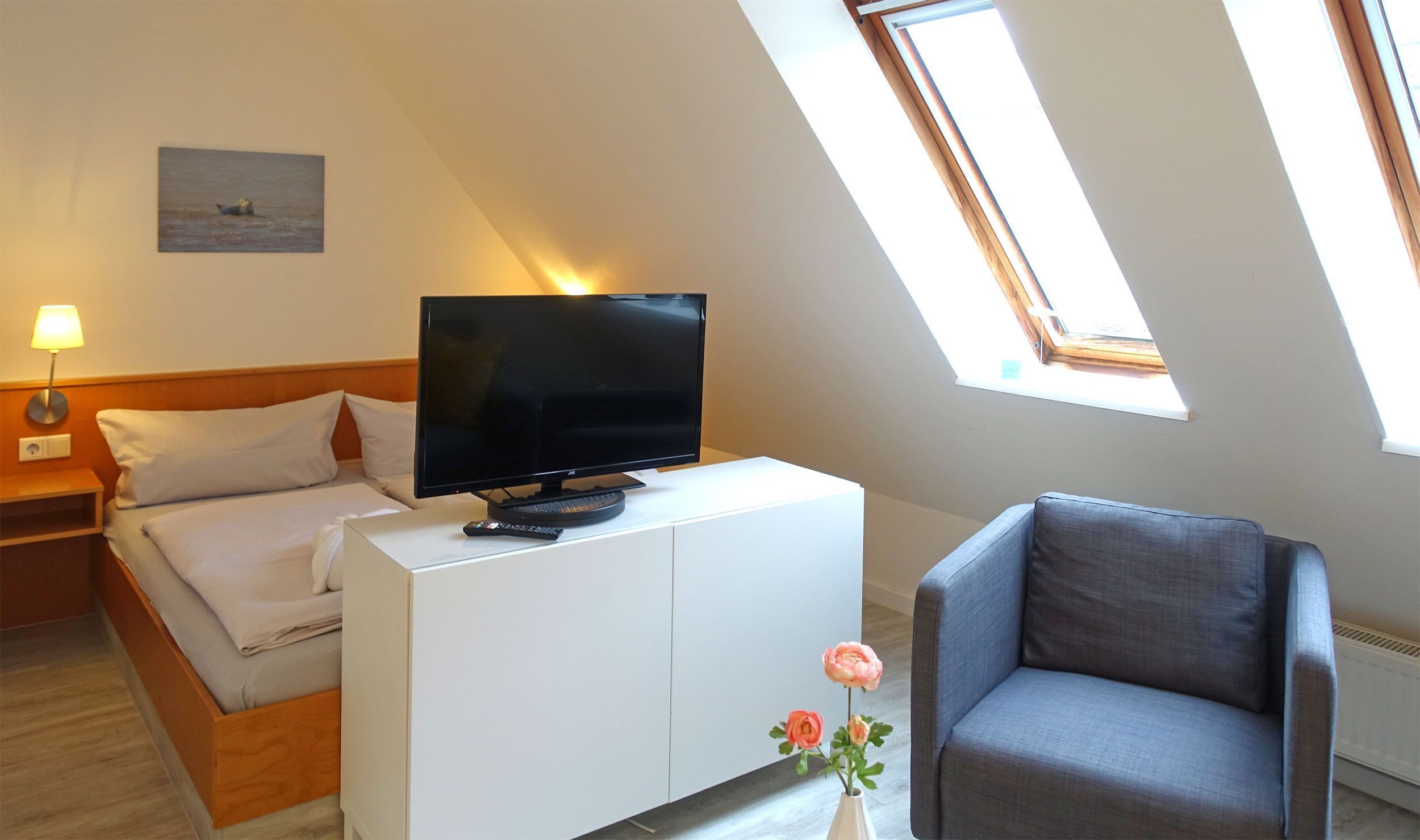 Una habitación de hotel con cama, un televisor sobre un mueble blanco, un sillón gris y un tragaluz.