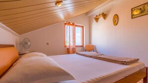Ein Schlafzimmer mit Holzboden, einem Bett mit weißer Bettwäsche, einem Ventilator, einem Fenster mit Vorhängen und einem kleinen Tisch mit einem Stuhl.