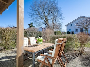 Un espace extérieur aménagé pour les repas, avec une table en bois et des chaises rembourrées, entouré de plantes et offrant une vue dégagée sur un immeuble d’habitation en arrière-plan.