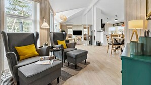 Un vaste espace de vie avec des canapés gris et des coussins jaunes, une table à manger et un coin cuisine équipé d’une télévision et d’une cheminée.