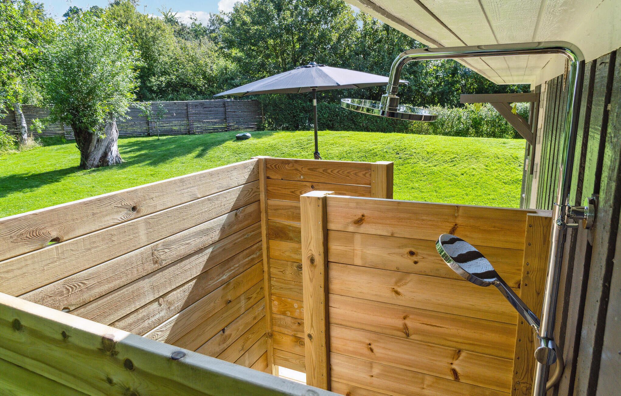 A wooden horse stall with a metal gate, a black umbrella, and a grassy yard with trees.