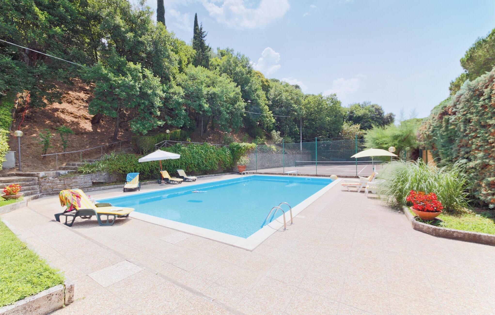 A rectangular swimming pool with a tiled deck, surrounded by a fence. There are lounge chairs and umbrellas along the poolside.