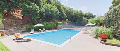 A rectangular swimming pool with a tiled deck, surrounded by a fence. There are lounge chairs and umbrellas along the poolside.