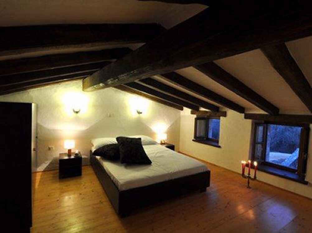 A bedroom with a wooden floor, a bed with white bedding, two bedside lamps, and a window with a view of the outside.