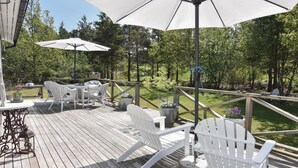A wooden deck with outdoor furniture, including white chairs and a table, surrounded by greenery.