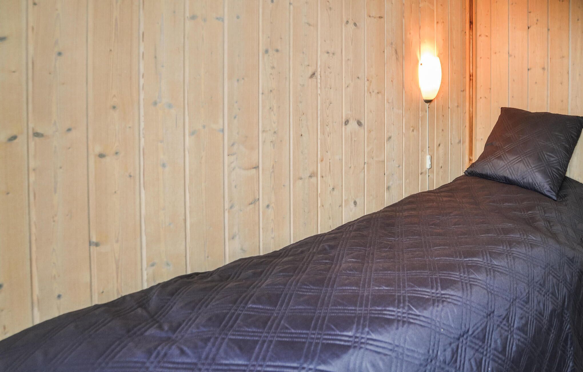 Un lit simple avec une couverture matelassée de couleur sombre et un oreiller assorti, le tout adossé à un mur en bois et éclairé par un luminaire fixé au mur.