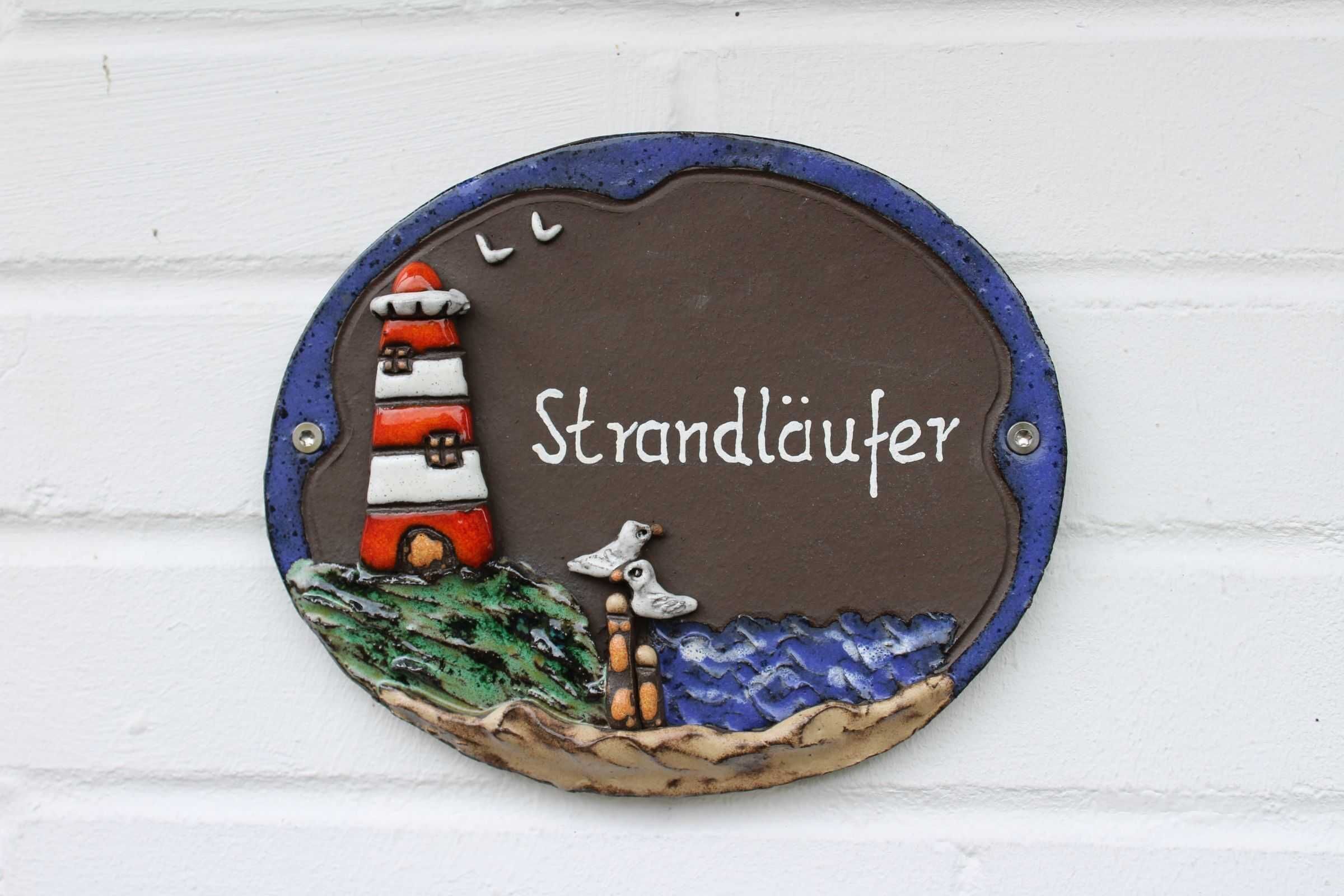 Ein rundes, metallenes Schild mit einer Leuchtturm-Illustration und dem Schriftzug „Strandlauer“ an einer weißen Ziegelwand.