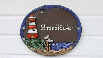 Ein rundes, metallenes Schild mit einer Leuchtturm-Illustration und dem Schriftzug „Strandlauer“ an einer weißen Ziegelwand.