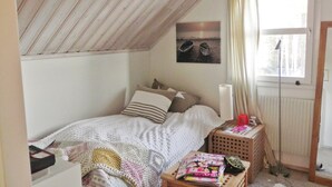 Un dormitorio con techo inclinado, una cama con almohadas, una mesita de noche con lámpara, una ventana con cortinas y un radiador.