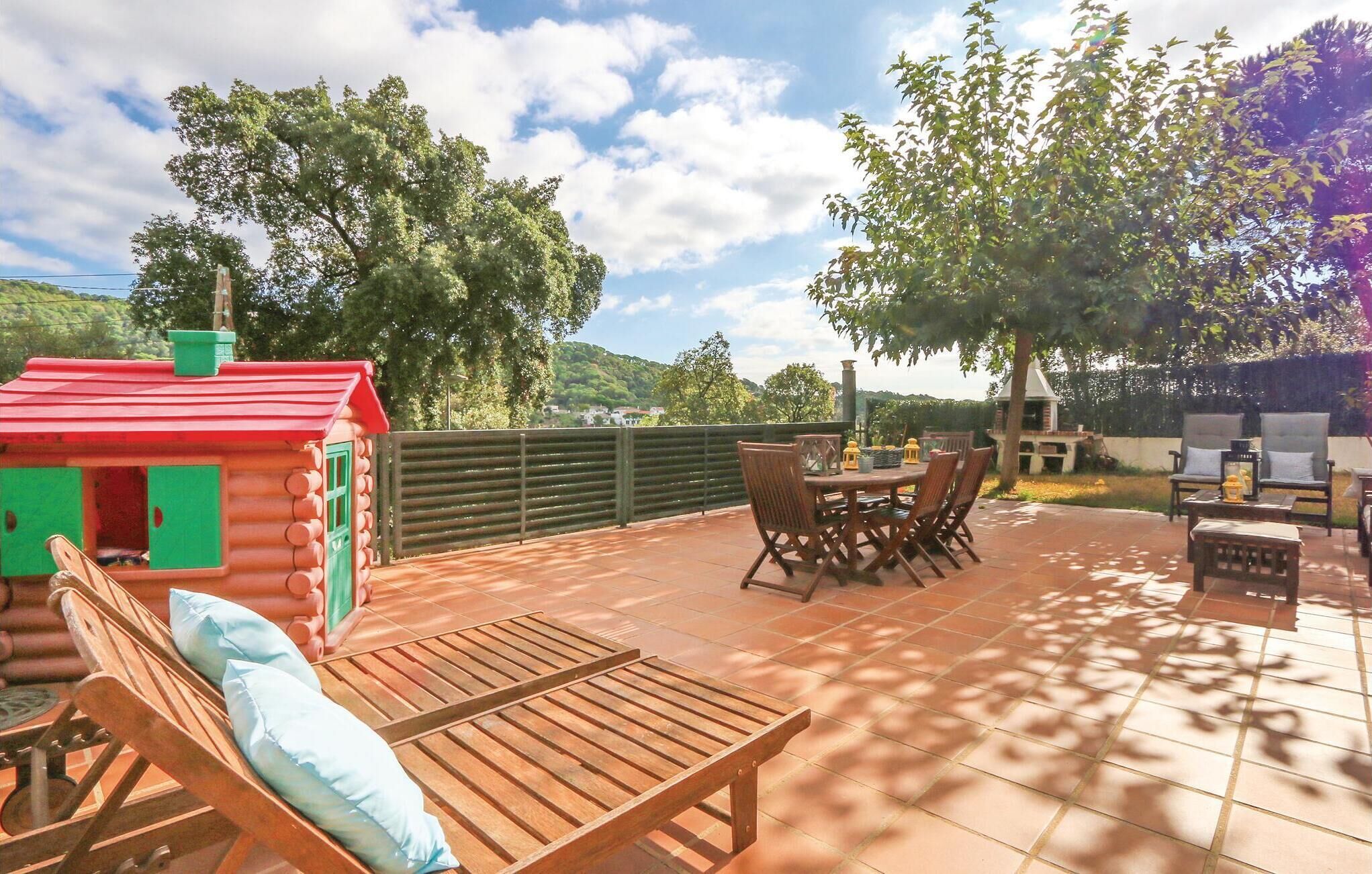 A patio with a wooden table and chairs, a small wooden cabin, and a view of trees and hills.