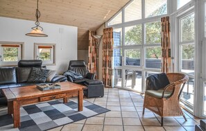 A spacious living room with a wooden ceiling, large windows, a black leather sofa, a grey armchair, a wicker chair, and a wooden coffee table. - Cozy home in Blåvand with sauna (Blåvand)